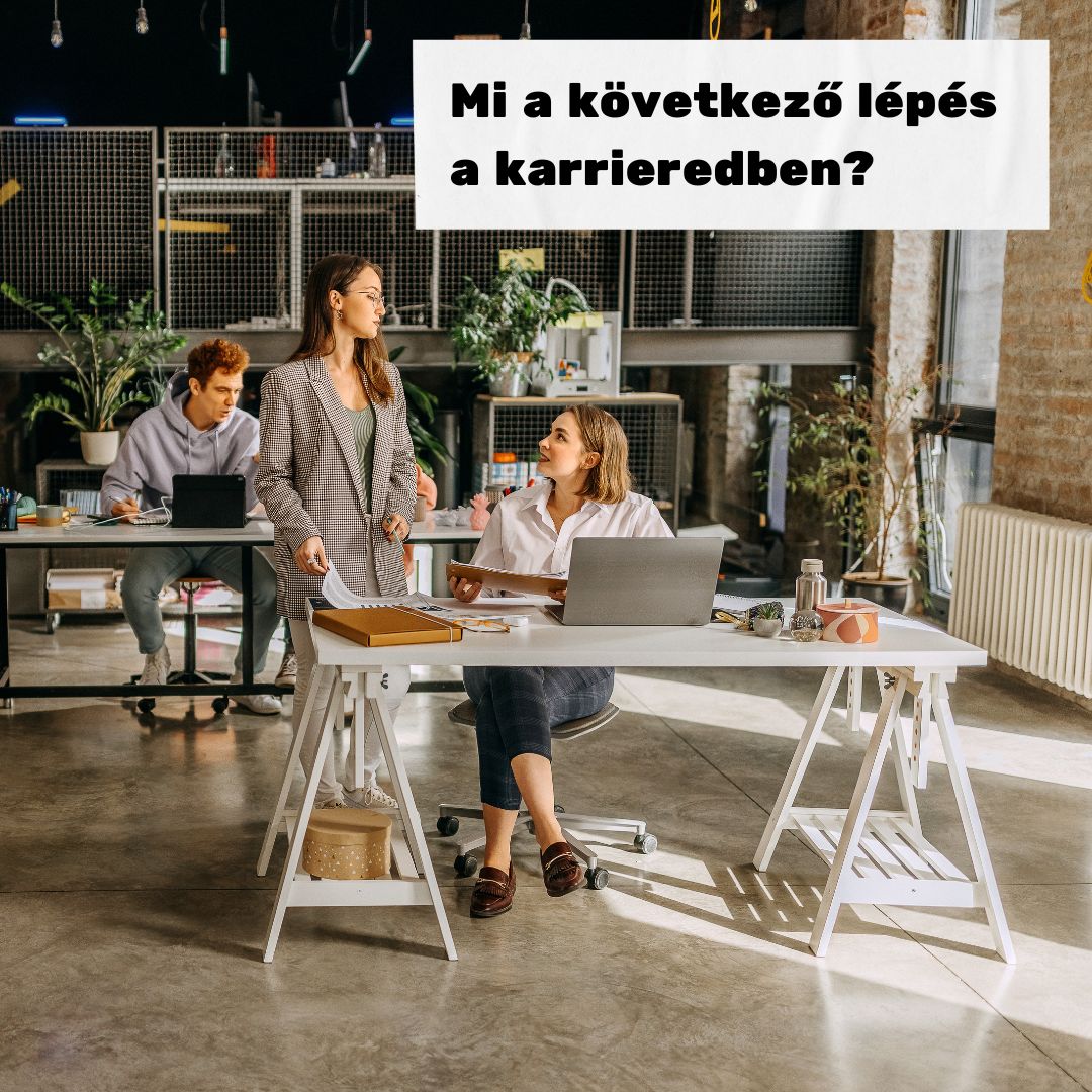 A képen három személy látható. Az előtérben két nő van. Az egyik személy áll, a másik ül. A háttérben egy férfi látható, aki dolgozik.
